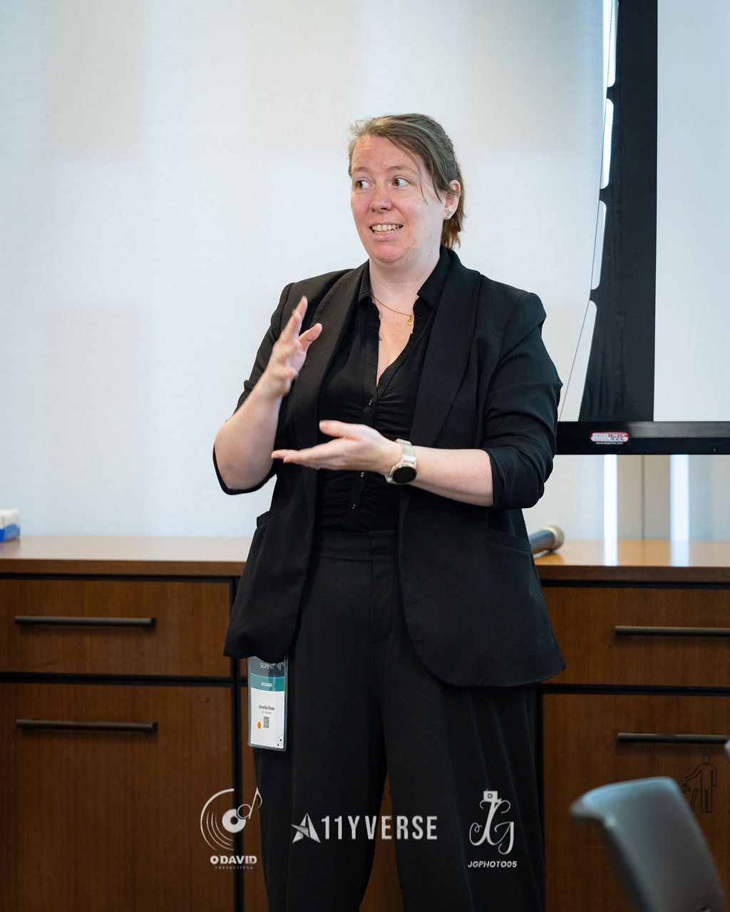 A person stands and gestures with both hands while speaking to a room, an attendee badge on a lanyard around their neck.