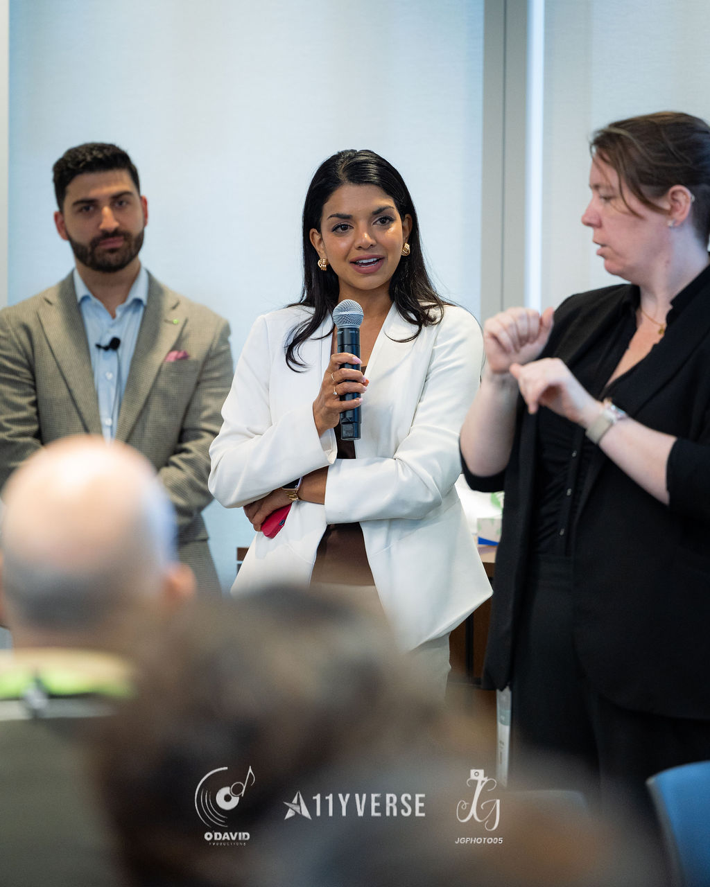 A person in a white blazer speaks into a handheld microphone while standing between two others, and a sign language interpreter signs to the side as the audience listens.