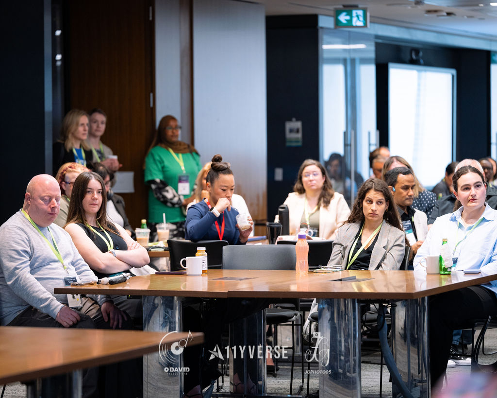 Rows of attendees seated at tables listening during a session, with drinks and cups on the tables and an exit sign overhead.