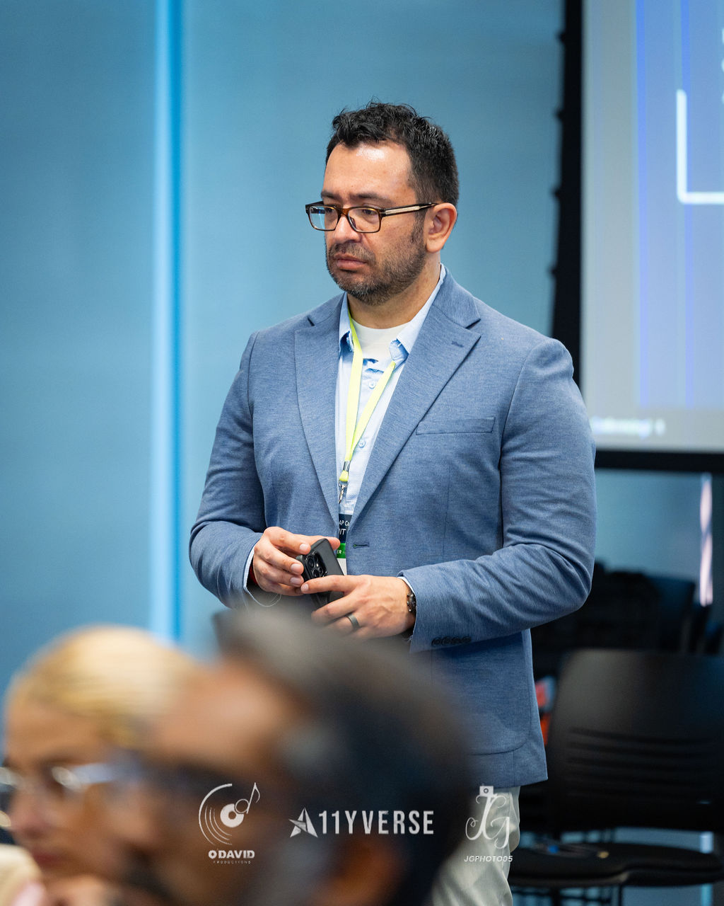 A person in a blazer and lanyard stands holding a presentation remote, looking down, with attendees seated in the foreground.