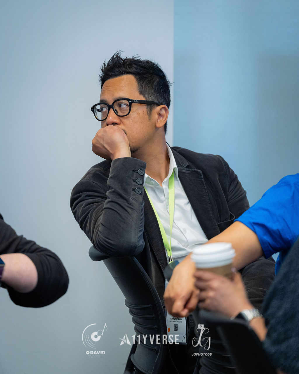 Close portrait of an attendee in a blazer and glasses resting their chin on a hand while listening, a takeaway coffee cup in the foreground.