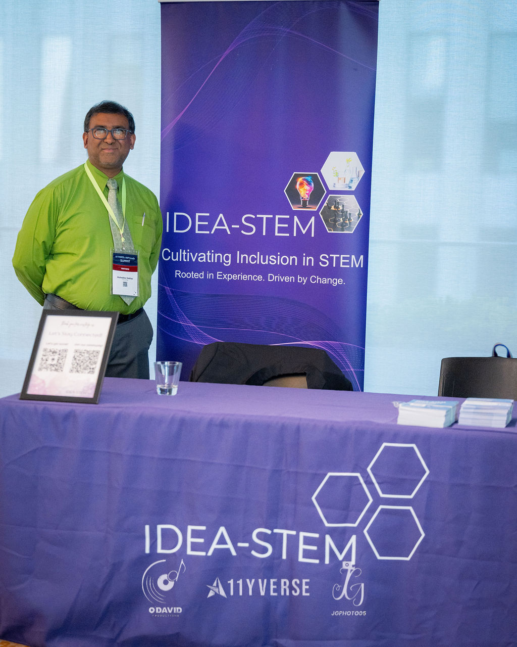 A person stands at an exhibitor booth behind a purple table, beside a banner for IDEA-STEM reading cultivating inclusion in STEM, with a QR code stand and printed cards on the table.