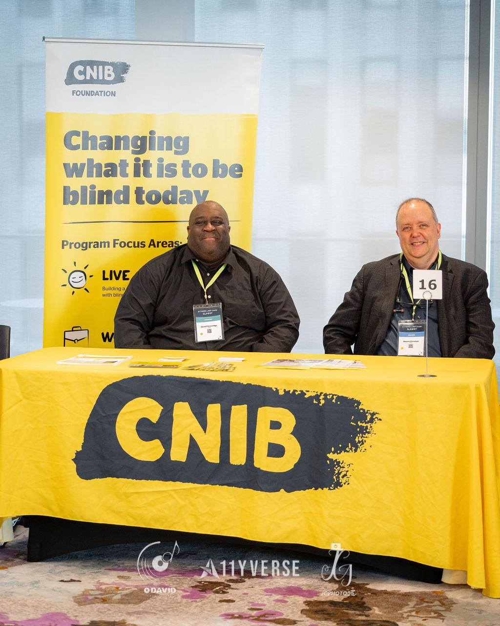 Two people sit smiling behind a CNIB Foundation booth with a bright yellow cloth and a banner about changing what it is to be blind, with printed materials laid out on the table.
