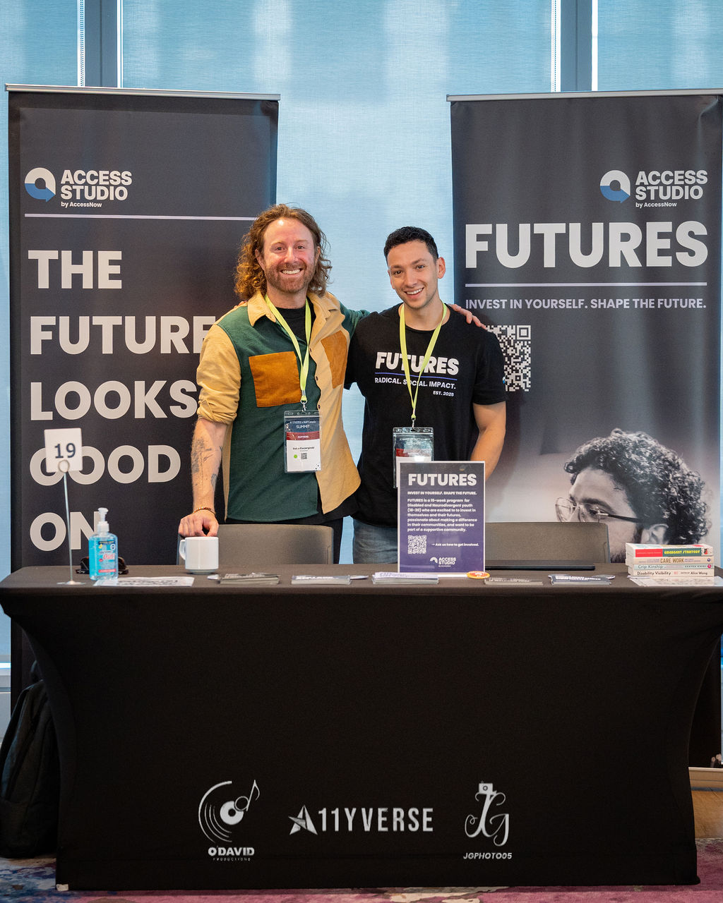 Two people pose smiling behind the Access Studio and Futures booth, the draped table holding printed materials in front of them.