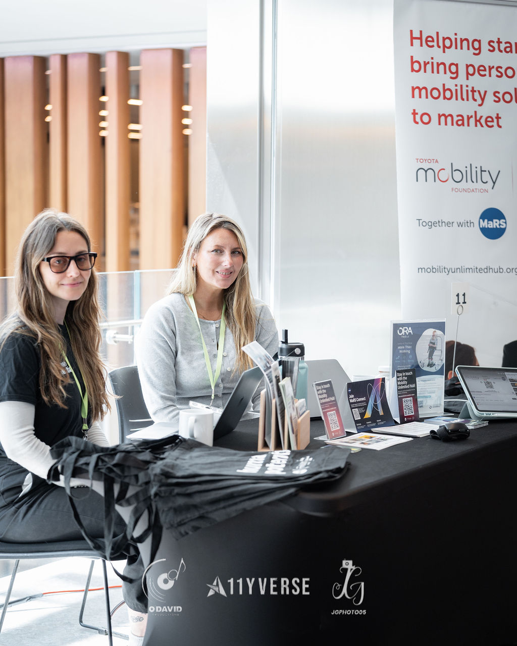 Two people sit smiling behind a Toyota Mobility Foundation booth, beside a banner about bringing personal mobility solutions to market, with brochures and a payment device on the table.