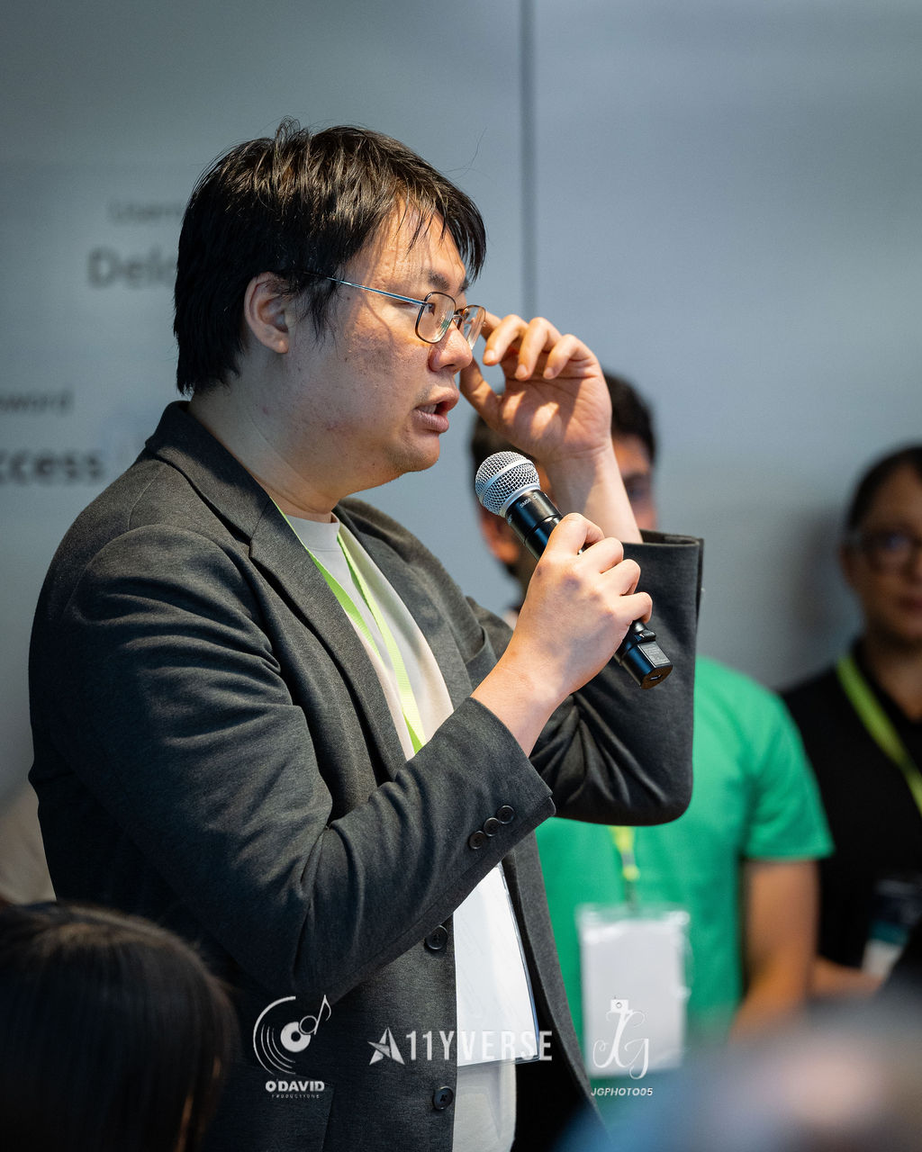 A person stands in the audience speaking into a handheld microphone to ask a question, with others seated and standing nearby and a presentation screen behind.