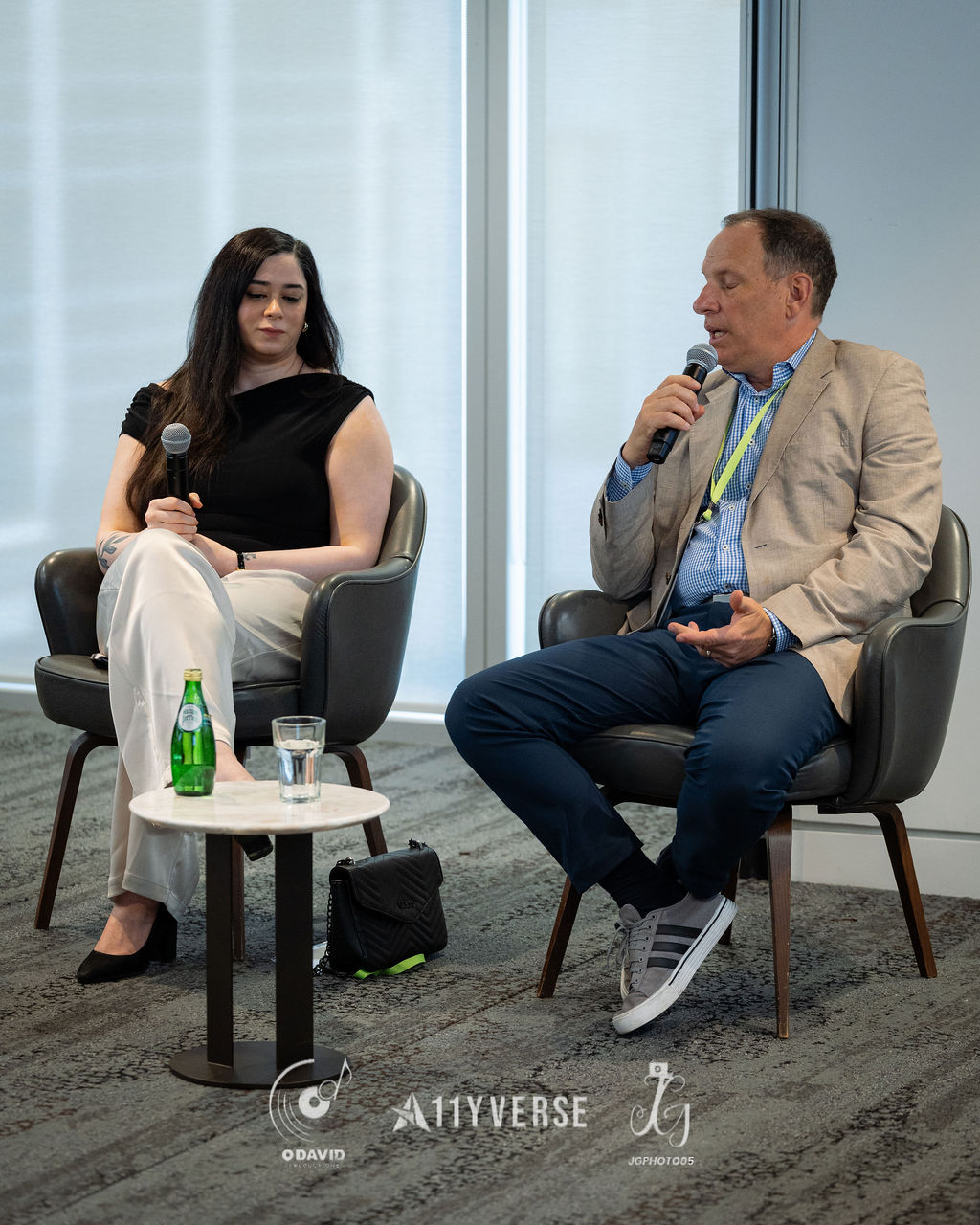 Two panelists sit in armchairs on stage, one speaking into a handheld microphone and the other listening, with a small side table of water between them.