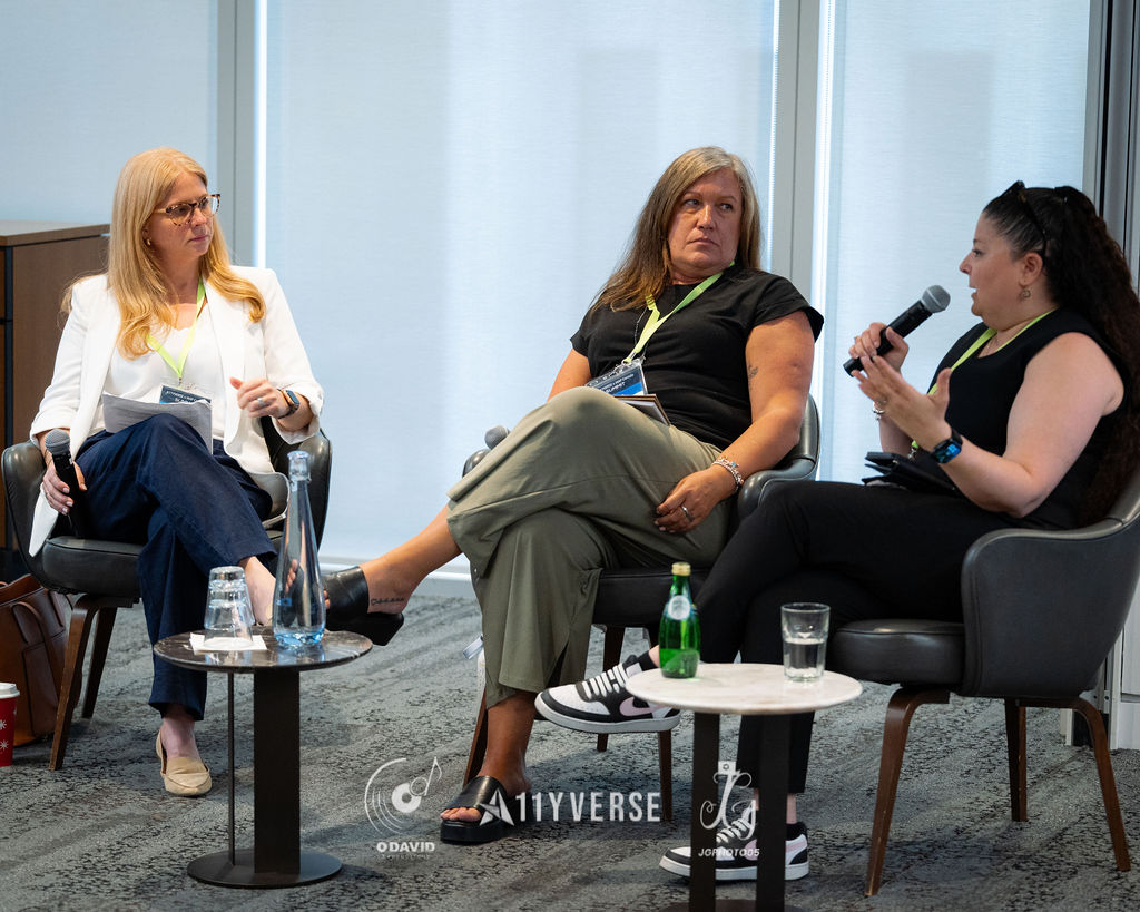 Three panelists sit in armchairs in a discussion, one speaking into a handheld microphone while the others listen, with side tables of water.