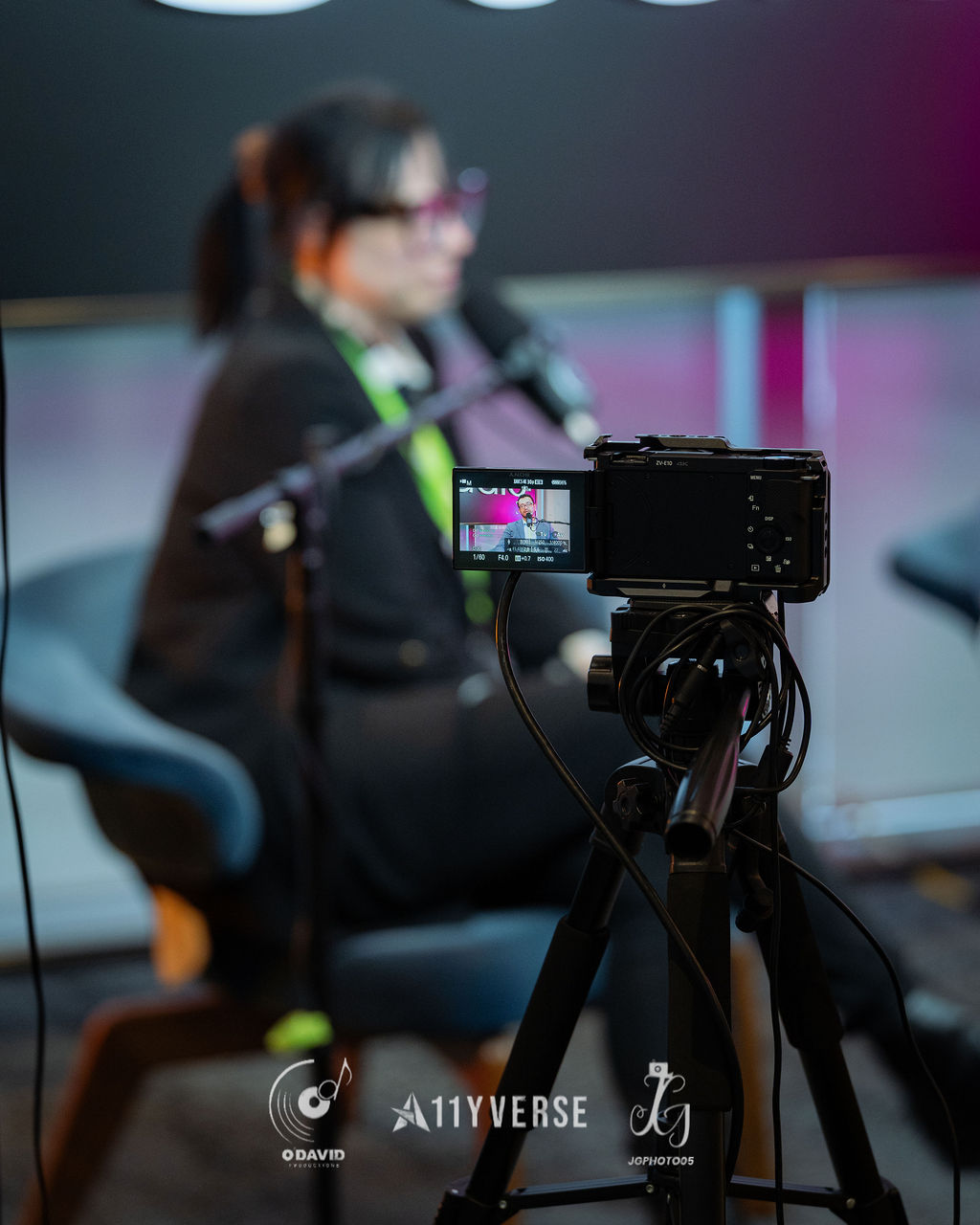 A video camera on a tripod films a seated panelist, who is softly blurred behind, the framed shot visible on the camera's screen.