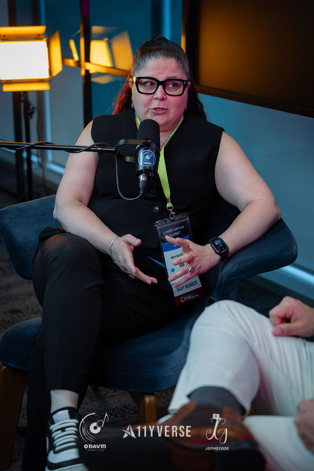 A person sits in an armchair gesturing while speaking into a podcast microphone, their lanyard showing an IAAP member credential, with studio lights behind.