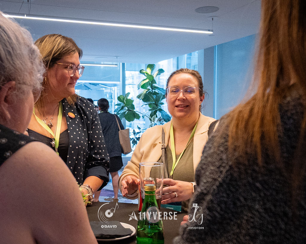 A small group of attendees stand in conversation and smile around a high table during a break, a tall plant and window behind them.