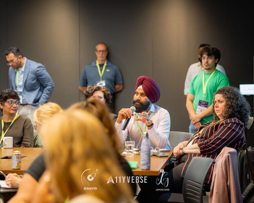 A person sits among attendees and speaks into a handheld microphone to ask a question, with others seated and standing around the tables listening.