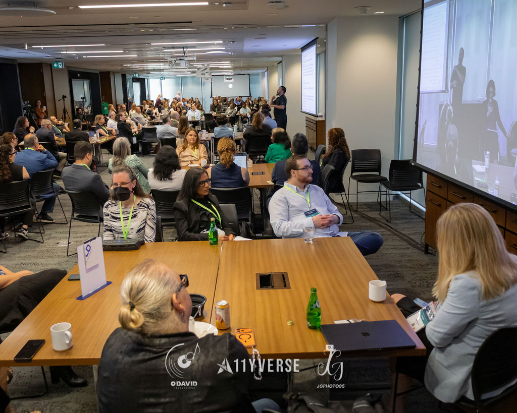 A wide view of a full session room with attendees seated at tables watching a presenter at the front, while a large screen on the right shows a live video feed of the speaker.